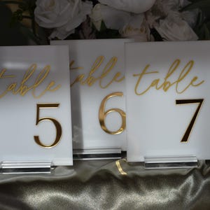 Pu&ograve; includere: Tre segnaposto bianchi con numeri da tavolo con lettere e numeri dorati. La parola "table" &egrave; scritta in corsivo sopra i numeri 5, 6 e 7. Ogni segnaposto &egrave; esposto su una base in acrilico trasparente.