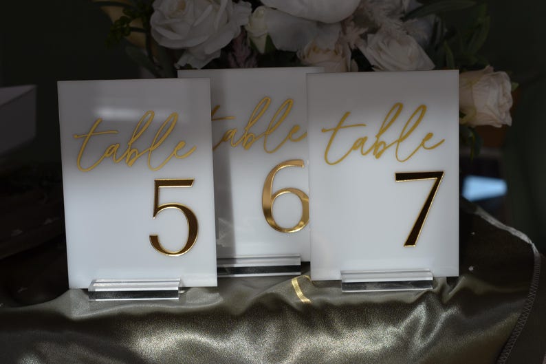 Pu&ograve; includere: Tre segnaposto bianchi con numeri da tavolo con lettere e numeri dorati. La parola "table" &egrave; scritta in corsivo sopra i numeri 5, 6 e 7. Ogni segnaposto &egrave; esposto su una base in acrilico trasparente.