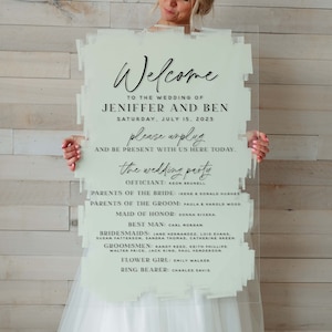 May include: A white acrylic sign with a green wash background and black text announcing a wedding for Jennifer and Ben on July 15, 2023. The sign lists the wedding party, including the officiant, parents of the bride and groom, maid of honor, best man, bridesmaids, groomsmen, flower girl, and ring bearer.