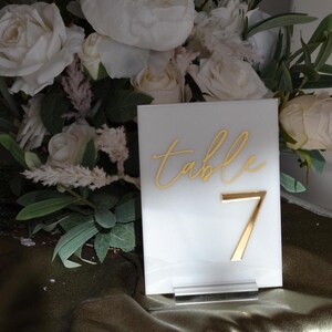 Pu&ograve; includere: Un segnaposto in acrilico bianco con scritte dorate e il numero 7. La parola "table" &egrave; scritta in corsivo. Il segnaposto &egrave; esposto davanti a un bouquet di rose bianche e verde.