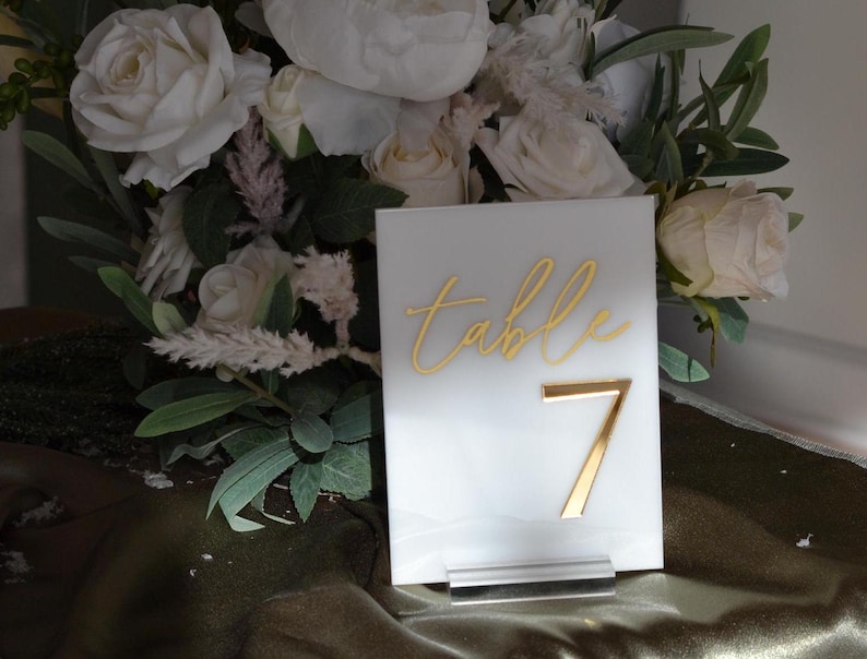 Pu&ograve; includere: Un segnaposto in acrilico bianco con scritte dorate e il numero 7. La parola "table" &egrave; scritta in corsivo. Il segnaposto &egrave; esposto davanti a un bouquet di rose bianche e verde.