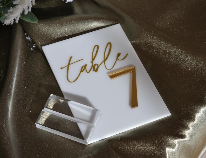 Pu&ograve; includere: Numero tavolo in acrilico bianco con testo dorato "table" e numero 7 dorato. Include due supporti in acrilico trasparente. Ideale per matrimoni ed eventi.