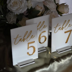 Pu&ograve; includere: Tre segnaposto bianchi con numeri da tavolo con lettere e numeri dorati. I segnaposto riportano la scritta "tavolo" seguita dai numeri 5, 6 e 7. Ogni segnaposto ha una base in acrilico trasparente. Lo sfondo comprende fiori bianchi e una tovaglia verde.