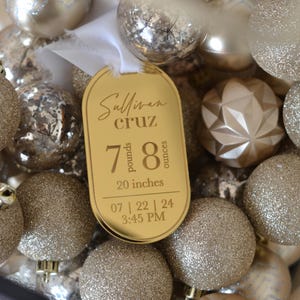 May include: A gold-coloured baby announcement bauble with the text "Sullivan Cruz" and the baby's birth stats: 7 pounds, 8 ounces, 20 inches, 07 | 22 | 24 3:45 PM. The bauble is surrounded by gold glitter Christmas decorations.