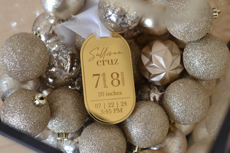May include: A gold-coloured baby announcement bauble with the text "Sullivan Cruz" and the baby's birth stats: 7 pounds, 8 ounces, 20 inches, 07 | 22 | 24 3:45 PM. The bauble is surrounded by gold glitter Christmas decorations.