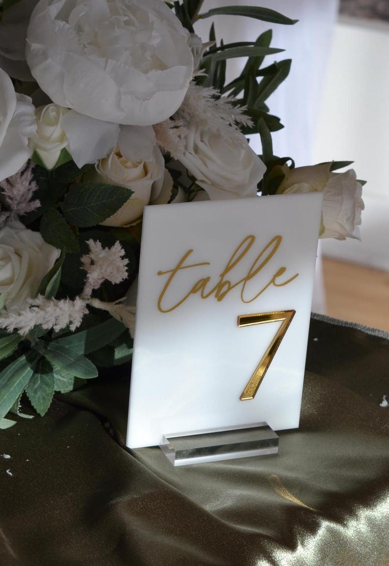 Pu&ograve; includere: Un segnaposto bianco con scritte dorate e il numero 7. Il segnaposto &egrave; esposto in un supporto in acrilico trasparente. Lo sfondo comprende fiori bianchi e verde, suggerendo un tema di matrimonio o evento.