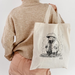 Puede incluir: Bolso tote de color crema con una ilustración en blanco y negro de una morsa bajo un paraguas bajo la lluvia. El bolso tiene asas largas y el texto "L'Atelier YACK". El bolso está hecho de tela resistente.
