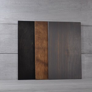 May include: Three rectangular wooden boards in varying shades of color. The boards are arranged vertically, with a black board on the left, a medium brown board in the middle, and a dark gray board on the right. The wood grain is visible.