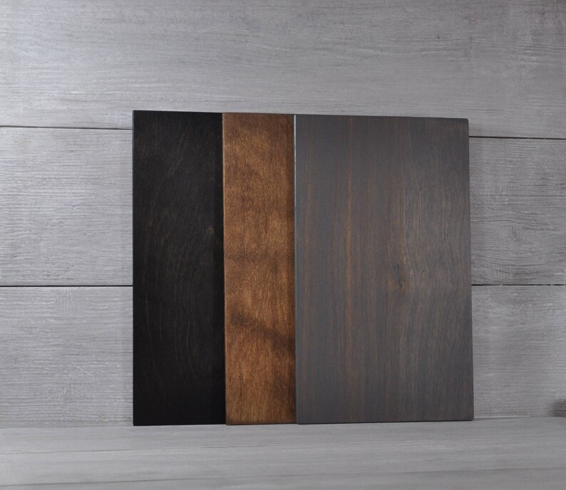 May include: Three rectangular wooden boards in varying shades of color. The boards are arranged vertically, with a black board on the left, a medium brown board in the middle, and a dark gray board on the right. The wood grain is visible.