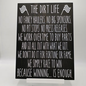 May include: A black wooden sign with white text that reads "THE DIRT LIFE" and other racing-related phrases. The sign has two checkered flags at the top. The sign is displayed on a small white easel.