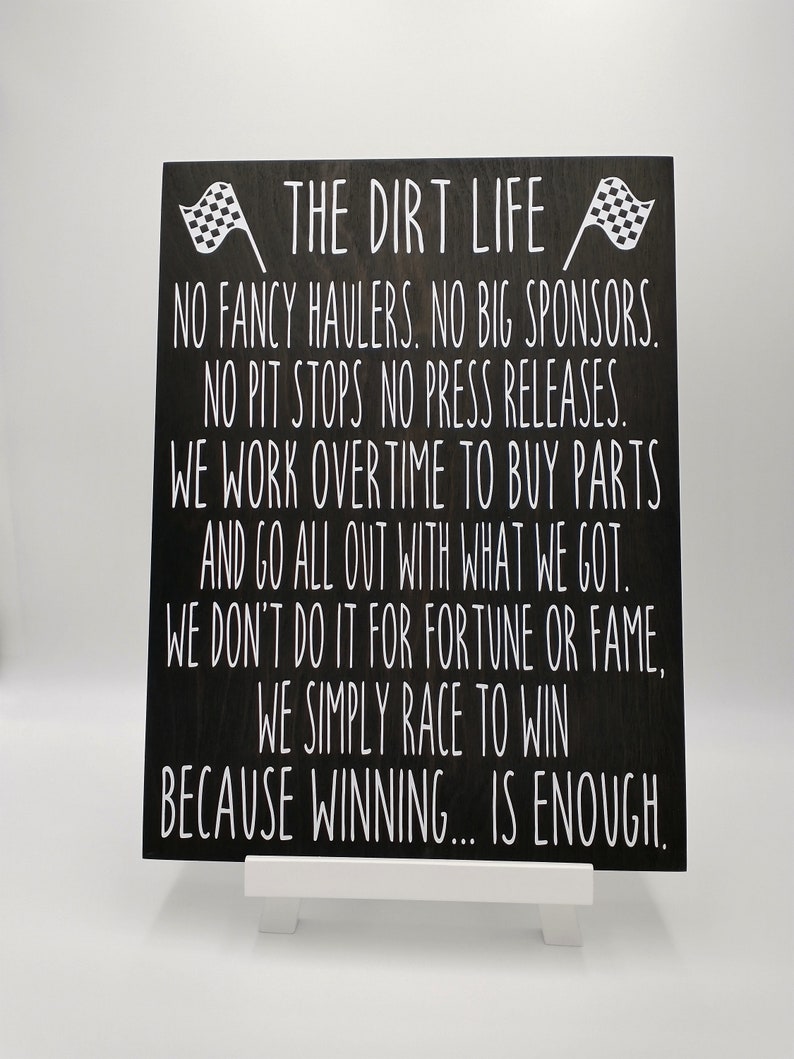 May include: A black wooden sign with white text that reads "THE DIRT LIFE" and other racing-related phrases. The sign has two checkered flags at the top. The sign is displayed on a small white easel.