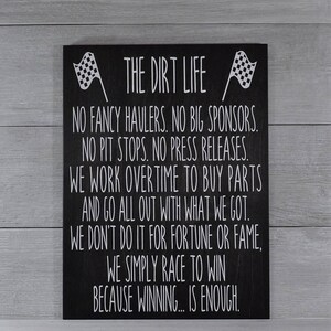 May include: Black wooden sign with white text and checkered flags. The sign reads "THE DIRT LIFE" and includes a motivational message about racing, including the phrase "WE SIMPLY RACE TO WIN BECAUSE WINNING... IS ENOUGH."
