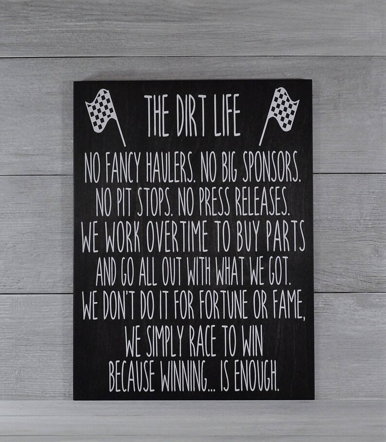 May include: Black wooden sign with white text and checkered flags. The sign reads "THE DIRT LIFE" and includes a motivational message about racing, including the phrase "WE SIMPLY RACE TO WIN BECAUSE WINNING... IS ENOUGH."