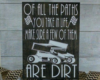 May include: A wooden sign with a black background and white text that reads "Of all the paths you take in life make sure a few of them are dirt". A white silhouette of a race car is in the centre of the sign.