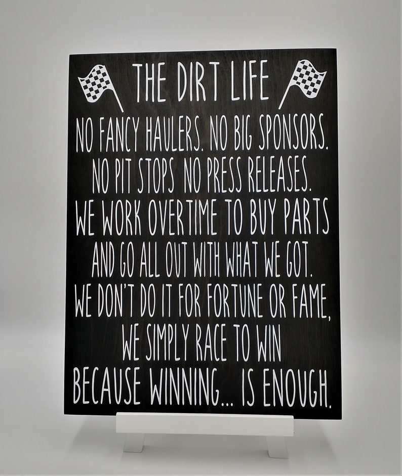 May include: A black wooden sign with white text that reads "THE DIRT LIFE." The sign includes the text "NO FANCY HAULERS, NO BIG SPONSORS." Two checkered flags are at the top. The sign is displayed on a white easel.