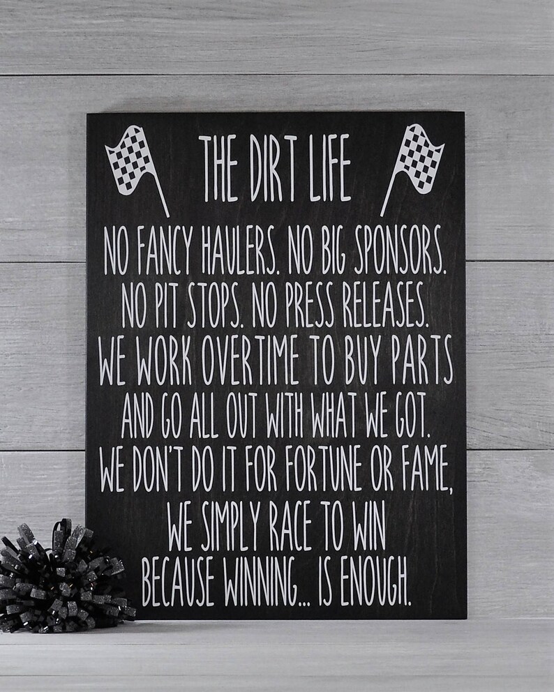 May include: Black wooden sign with white text that reads "THE DIRT LIFE" and a racing-themed message. Features two checkered flags. A decorative black object is in the lower left corner.