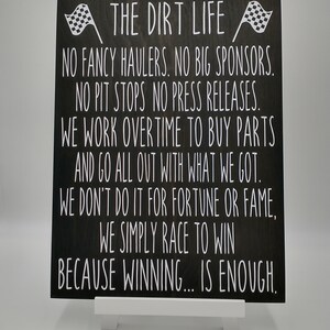 May include: A black wooden sign with white text and two checkered flags. The text reads "THE DIRT LIFE" followed by a motivational message about racing and winning. The sign is propped up on a white stand.