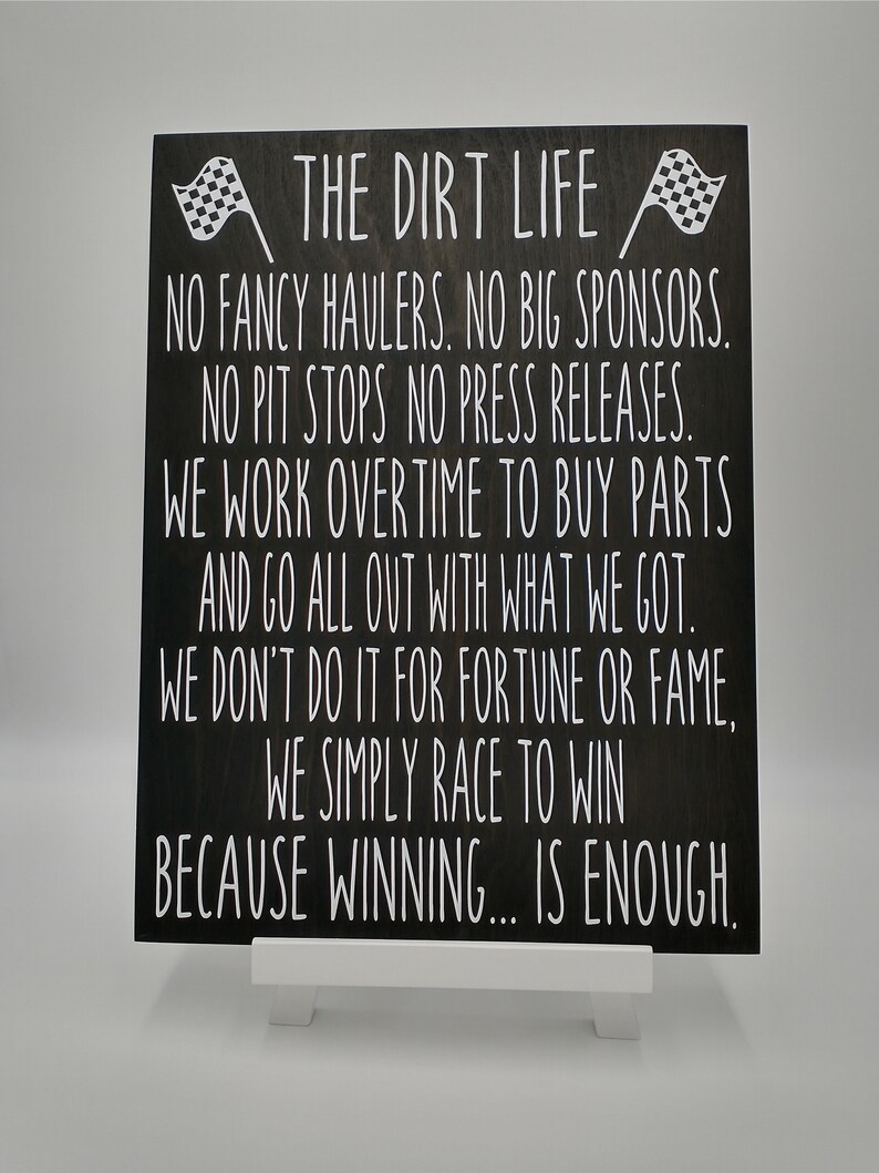 May include: A black wooden sign with white text and two checkered flags. The text reads "THE DIRT LIFE" followed by a motivational message about racing and winning. The sign is propped up on a white stand.