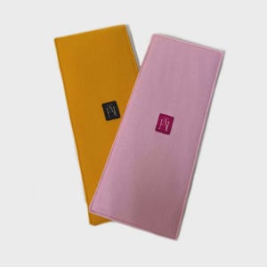 Op de afbeelding: Twee rechthoekige vilten items, één mosterdgeel en één roze, worden schuin weergegeven. Elk heeft een klein, vierkant label met een logo. De items lijken accessoires of organizers te zijn.