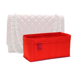 Op de afbeelding: Een felrode handtassenorganizer met meerdere compartimenten. De rechthoekige organizer is gemaakt van vilt. Een klein rood label met witte tekst is op de voorkant genaaid. Een witte handtas met een zilveren ketting staat op de achtergrond.