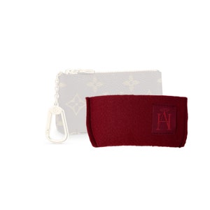 Può includere: Una piccola pochette con cerniera a zip di colore marrone e beige con un portachiavi argentato. La pochette è appoggiata su una pochette in feltro rosso con le lettere "AH" in un cerchio.
