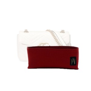 Peut inclure: Un organisateur de sac à main en feutre bordeaux, de forme rectangulaire, avec une petite étiquette noire portant du texte blanc. Un sac à main en cuir blanc matelassé avec une bandoulière en chaîne dorée est en arrière-plan.