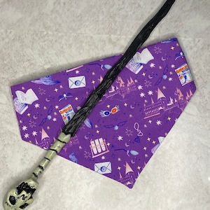 May include: A purple bandana with a white background and a variety of Harry Potter themed illustrations, including a Hogwarts castle, a golden snitch, a letter, and a pair of glasses. A black and white skull-shaped wand is lying on top of the bandana.