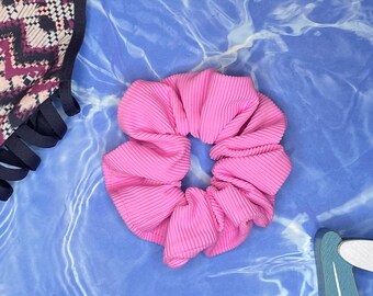 May include: A bright pink scrunchie with a ribbed texture sits on a blue and white patterned surface. A patterned swimsuit top and a blue starfish are also visible.