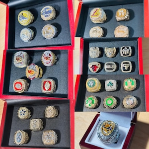 May include: Multiple championship rings displayed in red boxes. The rings are gold and silver, with various team logos and intricate designs. Some rings feature diamonds and colorful accents. The rings are arranged on a black surface.