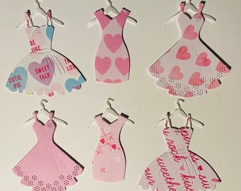 May include: Six paper cutouts of pink dresses on white hangers. The dresses have different patterns, including hearts, polka dots, and a tic-tac-toe board. The text on the dresses includes "Sweet Talk", "Cutie Pie", "Be Mine", "You Rock", "Sweetheart", and "A Kiss".
