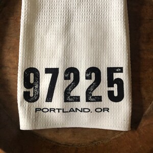 May include: White waffle weave kitchen towel with black text that reads "97225 PORTLAND, OR".
