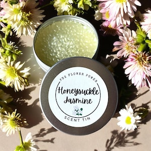 May include: A silver scent tin with the words "Honeysuckle Jasmine" and "PDX Flower Power" printed on a white label. The tin is open, revealing a light green, bead-like substance. The image is surrounded by white and pink flowers.