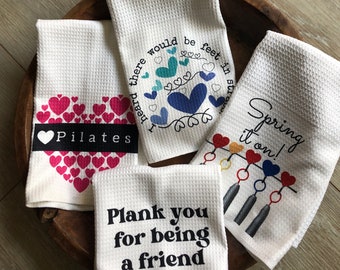 May include: Four white kitchen towels with different designs and sayings. One towel has a pink heart with the word "Pilates" in black text. Another towel has a blue heart with the text "I heard there would be feet in straps". A third towel has a colorful heart design with the text "Spring it on!" and a fourth towel has the text "Plank you for being a friend".