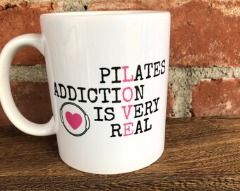 May include: White ceramic coffee mug with black text that reads "Pilates Addiction is Very Real" with a pink heart graphic.