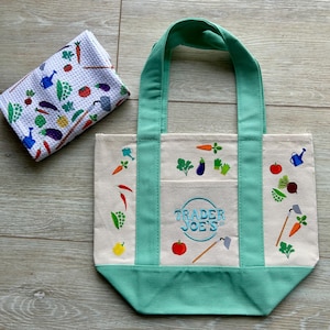 May include: A canvas tote bag with green accents and handles, featuring colorful illustrations of vegetables and gardening tools. The bag has the text "Trader Joe's" in a circular design. A matching patterned kitchen towel with vegetable illustrations is next to the bag.