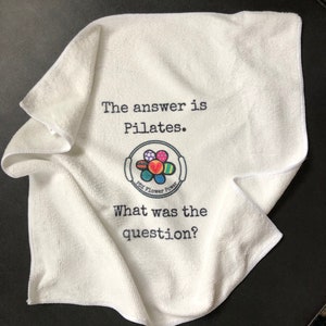 May include: White cotton towel with the text "The answer is Pilates." and a colorful flower logo with the text "Pix Flower Power". Below the logo is the text "What was the question?"