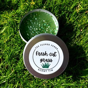 May include: A silver tin with a white label that reads "PDX FLOWER POWER Fresh cut grass SCENT TIN". The tin is filled with a green scented wax with glitter.