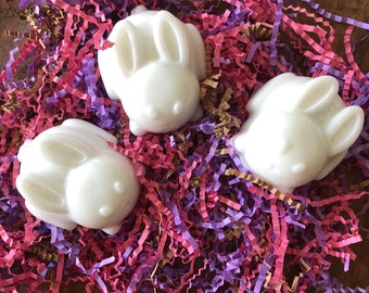 May include: Three white bunny-shaped soaps rest on a bed of colorful shredded paper. The soaps have detailed features, including ears, eyes, and a nose. The paper is a mix of pink, purple, and gold, creating a festive and spring-like scene.