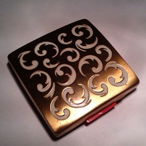 Op de afbeelding: Een vierkant, vintage compactpoederdoosje met een goudkleurige metalen afwerking. De bovenkant heeft een herhalend patroon van witte, gestileerde krullen. Een rode sluiting is zichtbaar aan de zijkant. Dit cosmetica-etui is een klassiek accessoire.