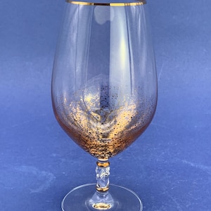 May include: A clear wine glass with a gold rim and gold glitter accents near the base. The stem has a decorative design with gold accents. The glass sits on a clear, round base.