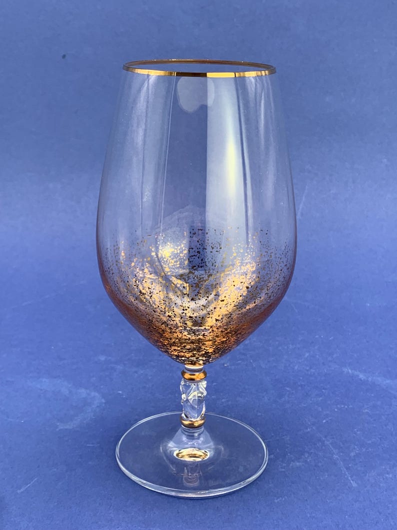 May include: A clear wine glass with a gold rim and gold glitter accents near the base. The stem has a decorative design with gold accents. The glass sits on a clear, round base.