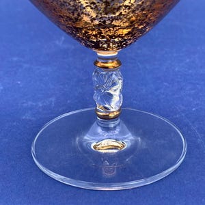 May include: A clear glass cocktail glass with a gold-colored rim and a gold and brown textured bowl. The stem has a clear, carved design with gold accents, and the base is a clear, flat circle. The glass is set against a blue background.