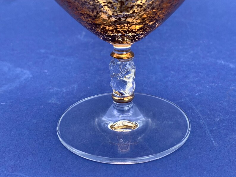 May include: A clear glass cocktail glass with a gold-colored rim and a gold and brown textured bowl. The stem has a clear, carved design with gold accents, and the base is a clear, flat circle. The glass is set against a blue background.