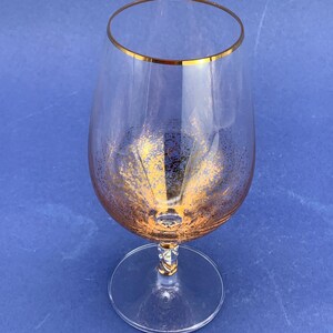 May include: A clear brandy snifter with a gold rim and gold flecks. The stem has a decorative design. The glass sits on a round base. The background is a solid blue.
