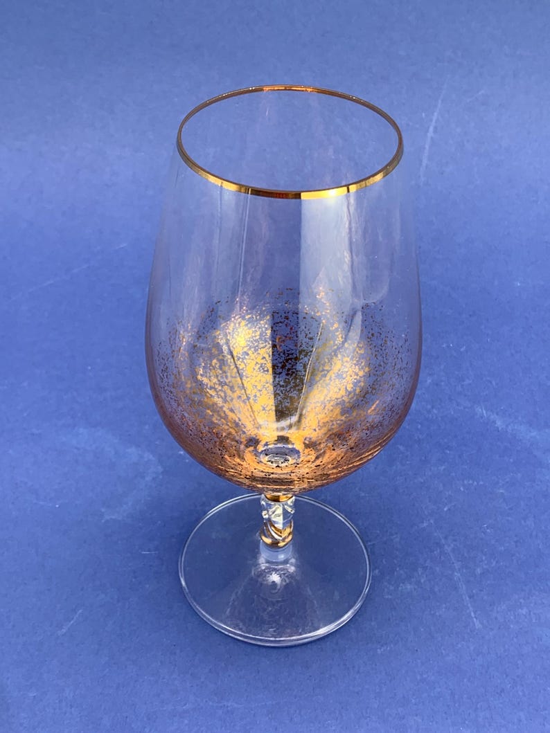 May include: A clear brandy snifter with a gold rim and gold flecks. The stem has a decorative design. The glass sits on a round base. The background is a solid blue.