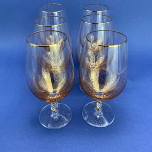 May include: Six clear wine glasses with gold rims and gold flecks inside. The glasses have a rounded bowl shape and sit on a clear stem and base. The glasses are arranged on a blue surface.