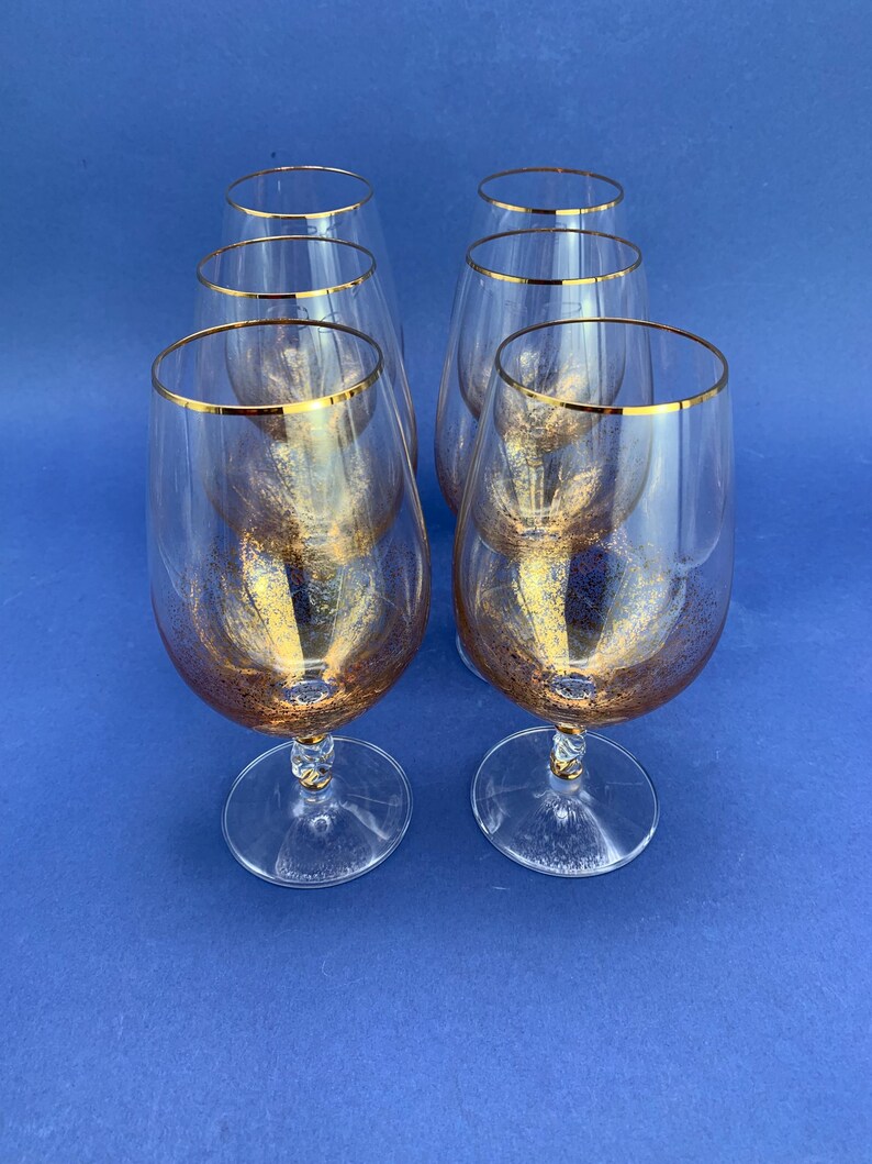 May include: Six clear wine glasses with gold rims and gold flecks inside. The glasses have a rounded bowl shape and sit on a clear stem and base. The glasses are arranged on a blue surface.