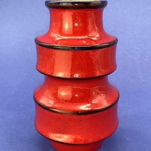 Puede incluir: Un jarrón de cerámica rojo con acabado brillante. El jarrón tiene forma cilíndrica con tres secciones distintas, cada una separada por una banda marrón oscuro. El borde superior también es marrón oscuro. Es un objeto decorativo.