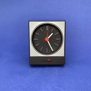 Pode incluir: Um relógio analógico preto e prateado com mostrador preto, ponteiros brancos e ponteiro de segundos vermelho. O relógio tem formato retangular e o texto "SOLARIS" está impresso na parte inferior.