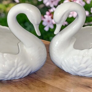 Può includere: Una coppia di ciotole in ceramica bianca a forma di cigno con dettagliate texture di piume modellate sull'esterno. Ogni cigno presenta un lungo collo ricurvo e una finitura liscia e lucida, con la sezione aperta che forma il corpo.